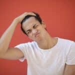 Young man making a funny face, scratching head against a red backdrop, showing playful expression.
